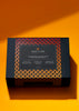 Black matte gift box with a black, yellow,  and red gradient belly band that reads "fruition chocolate: chocolate coated nuts gift collection". The box sits on an orange background