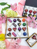 Black confection box containing 16 chocolate confections of various shapes and colors, surrounded by pink and yellow flowers- Fruition Chocolate