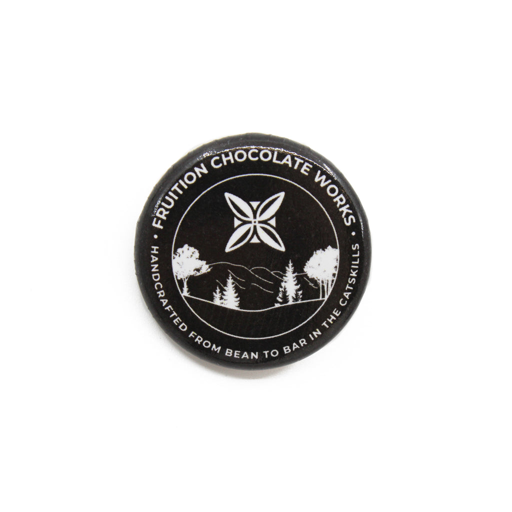 Round button style pin with a black background, and the text and image are white. Text around the outside says "fruition chocolate works, handcrafted from bean to bar in the catskills", and the center is a drawing of mountains and trees