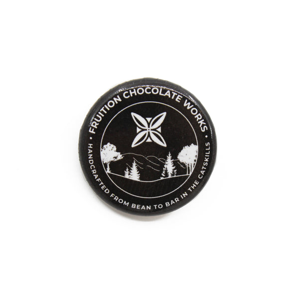 Round button style pin with a black background, and the text and image are white. Text around the outside says "fruition chocolate works, handcrafted from bean to bar in the catskills", and the center is a drawing of mountains and trees