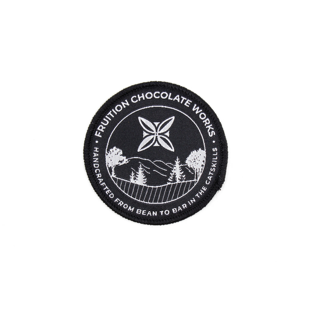 Black round woven patch with white text around the outside that reads "fruition chocolate works, handcrafted from bean to bar in the catskills". The center features the Fruition logo above a scene of trees and mountains