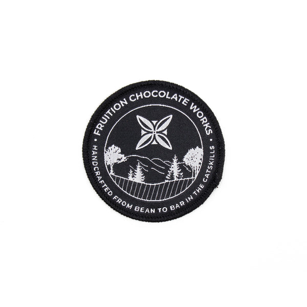 Black round woven patch with white text around the outside that reads "fruition chocolate works, handcrafted from bean to bar in the catskills". The center features the Fruition logo above a scene of trees and mountains